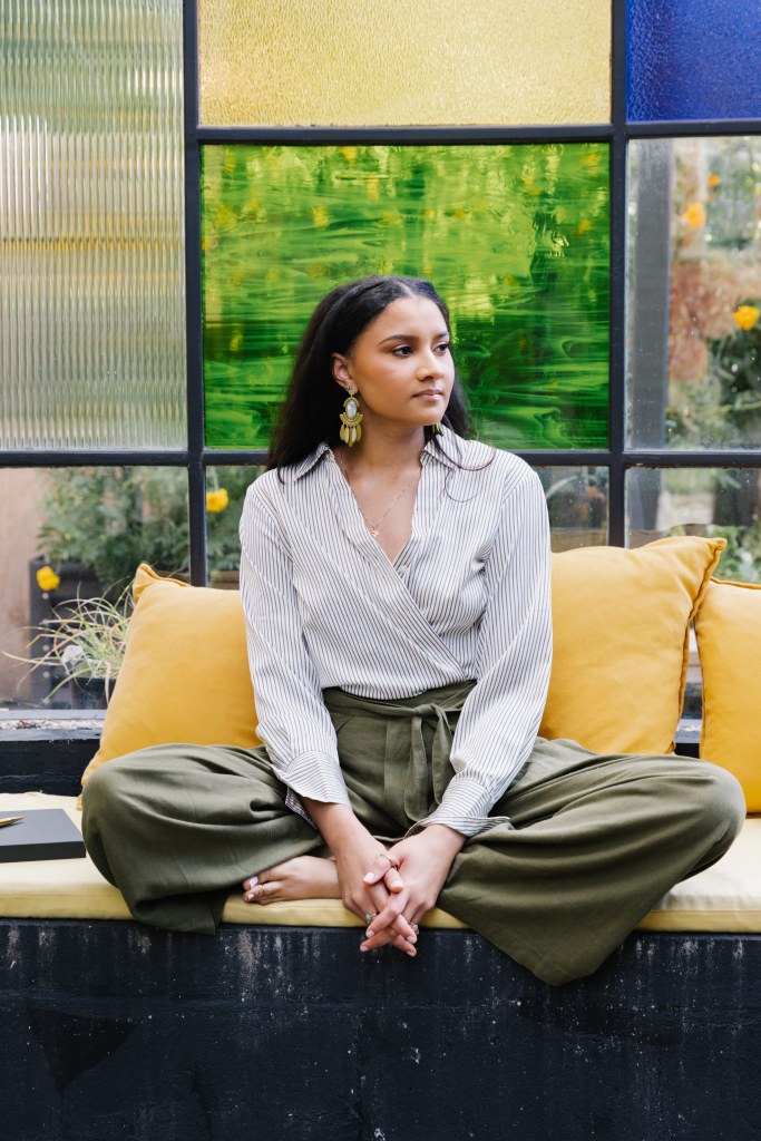 Victoria sitting cross-legged on a yellow bench in front of a mosaic stained glass window.