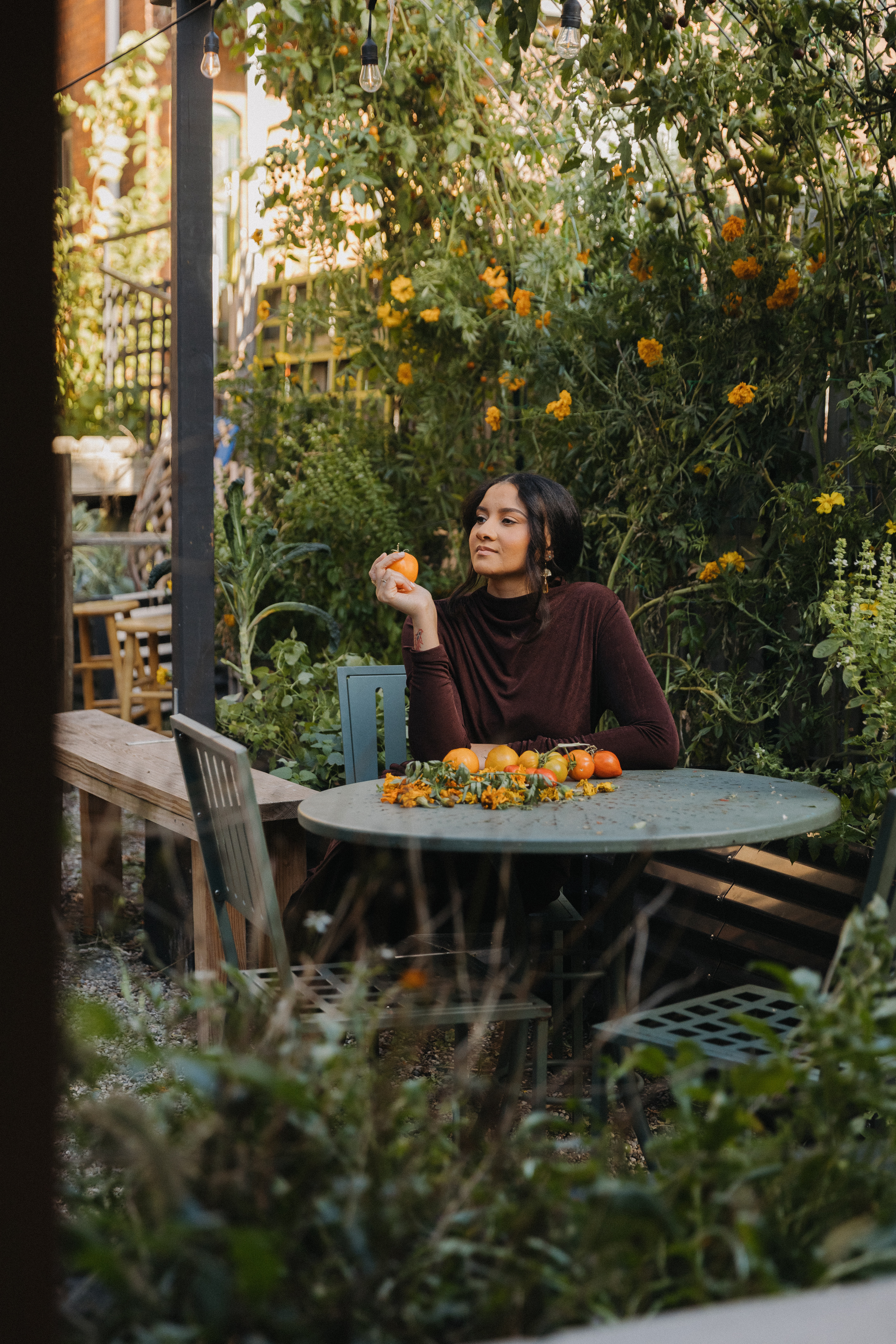 Victoria seated at a garden table with orange fruits and flowers, surrounded by lush greenery.