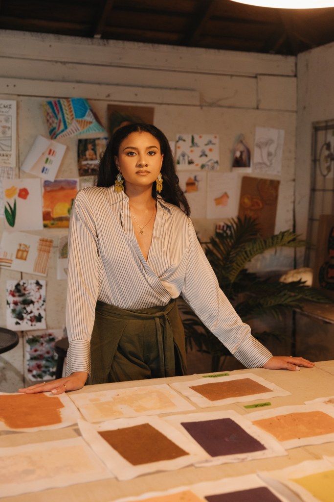 Victoria in an art studio standing at a table covered with art samples with artwork displayed on the wall.